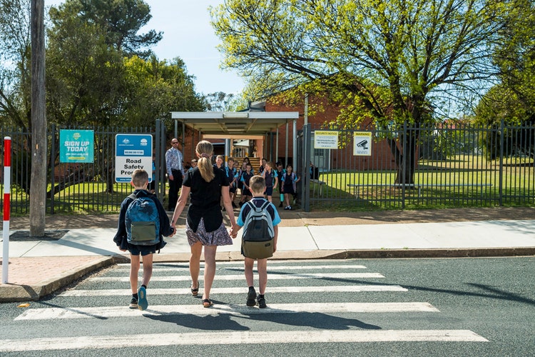 A mother walking her two children across the pedestrian crossing. The Principal and other students are waiting at the school gate.