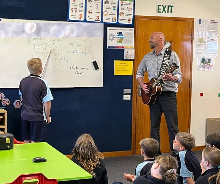The principal leads a music activity, playing the guitar while a student tracks the beat on the whiteboard.