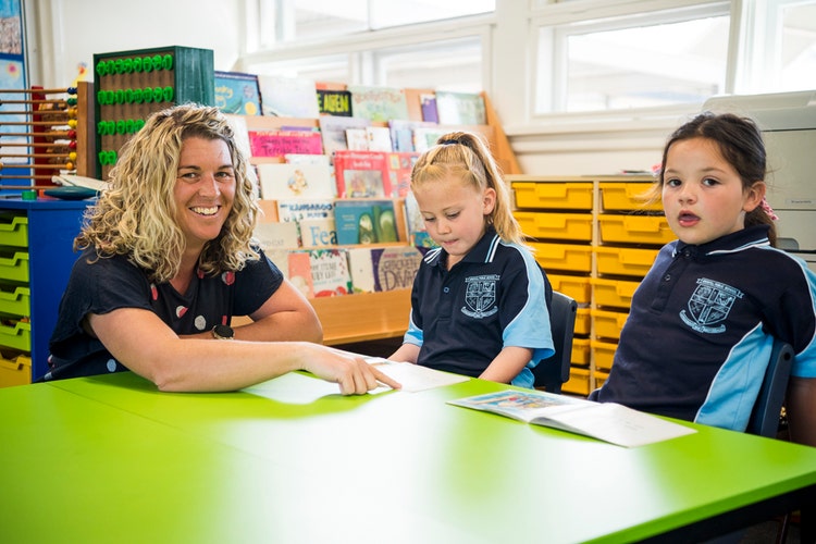 A teacher assists 2 students with their reading.